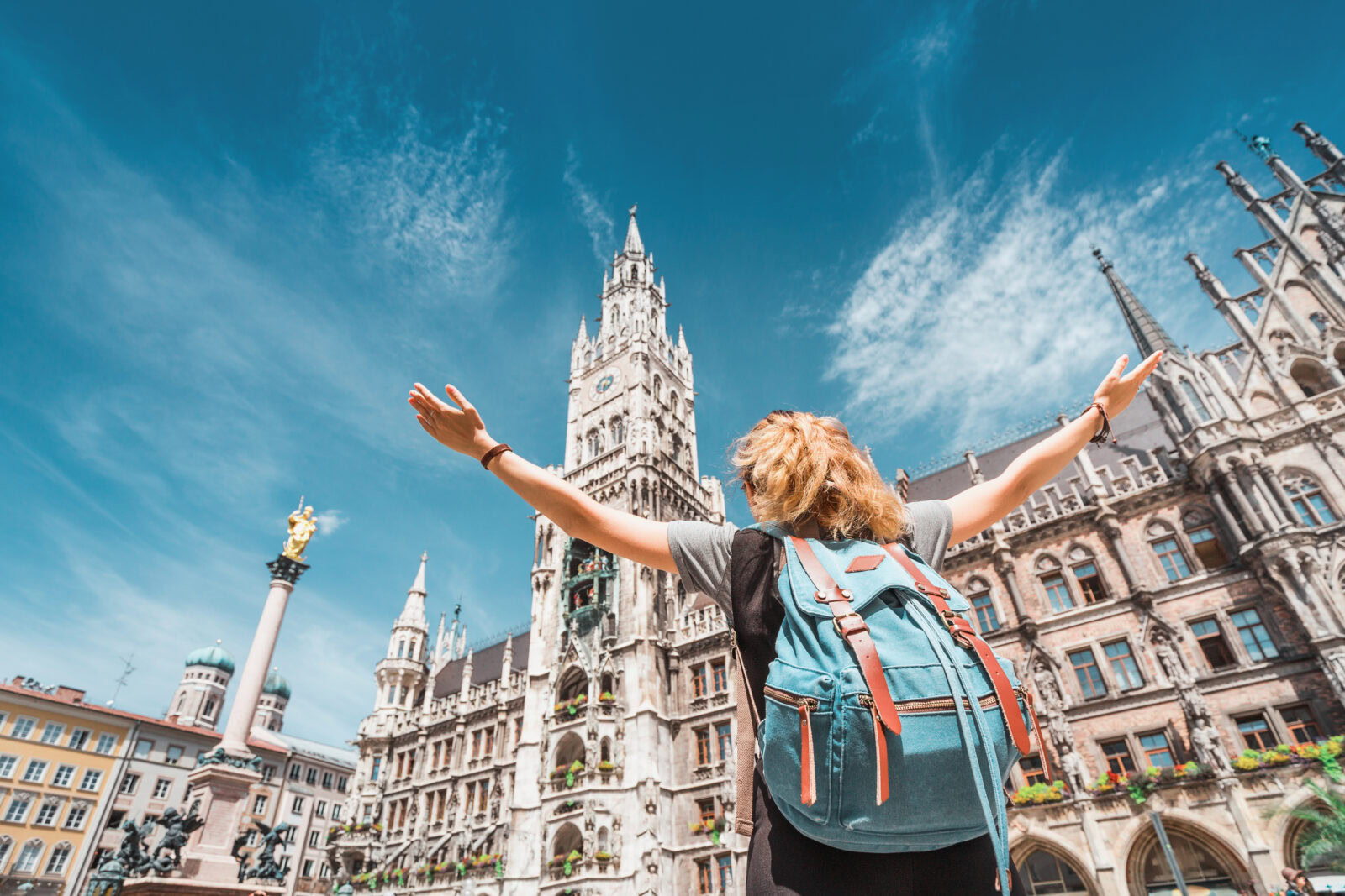 Studieren in München. Junge Studentin mit Rucksack steht mit ausgestreckten Armen vor dem Neuen Rathaus am Münchner Marienplatz.