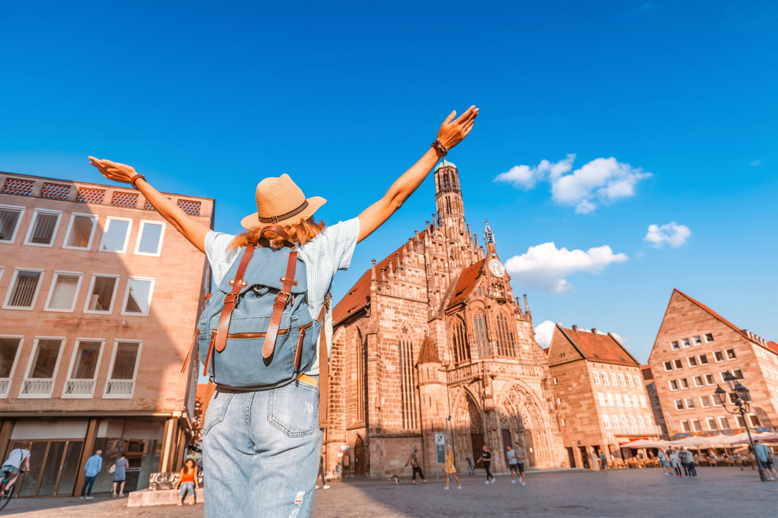 Young female student with a backpack and hat standing with outstretched arms in front of the St. Lorenz Church, symbolizing studying in Nuremberg